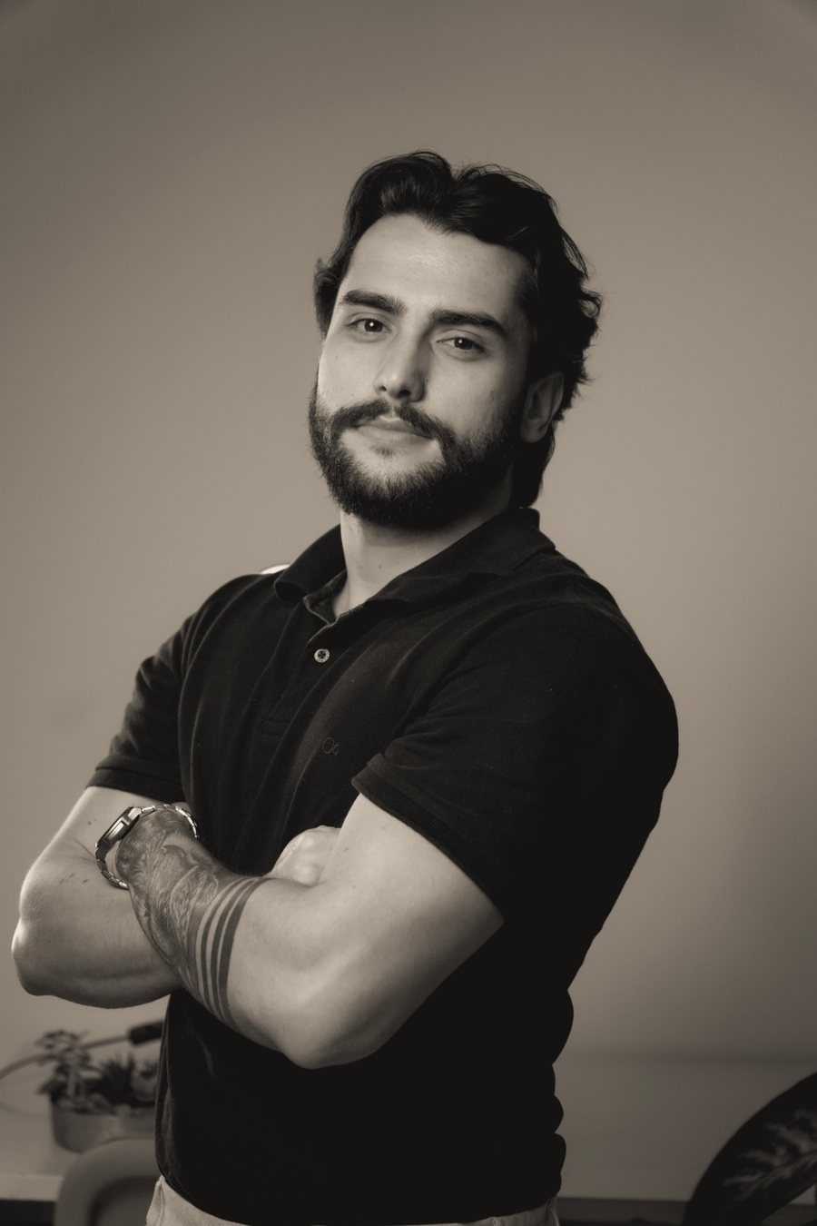 Thiago Maia, de barba e camisa polo preta, com os braços cruzados, em retrato em tom sépia